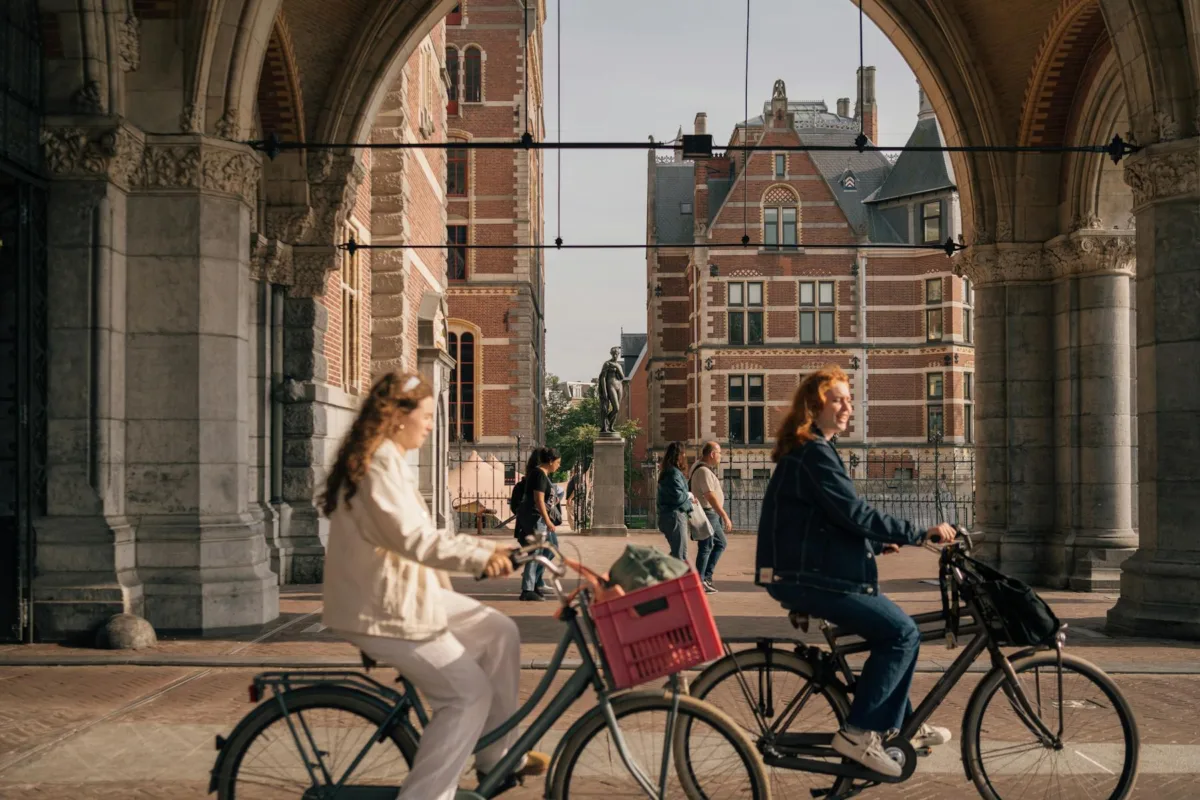 Warum sollten Sie in Amsterdam Urlaub machen? Frauen radeln unter einem Amsterdamer Bogen mit historischen Gebäuden im Hintergrund.